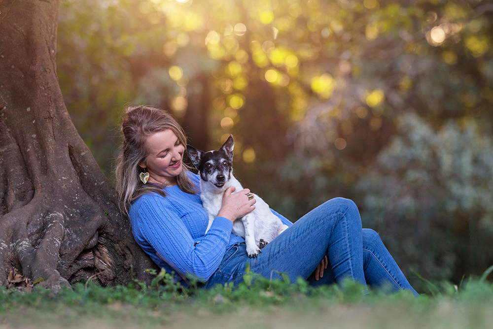 woman sitting against a tree with her fox terrier on her lap in the late afternoon golden light